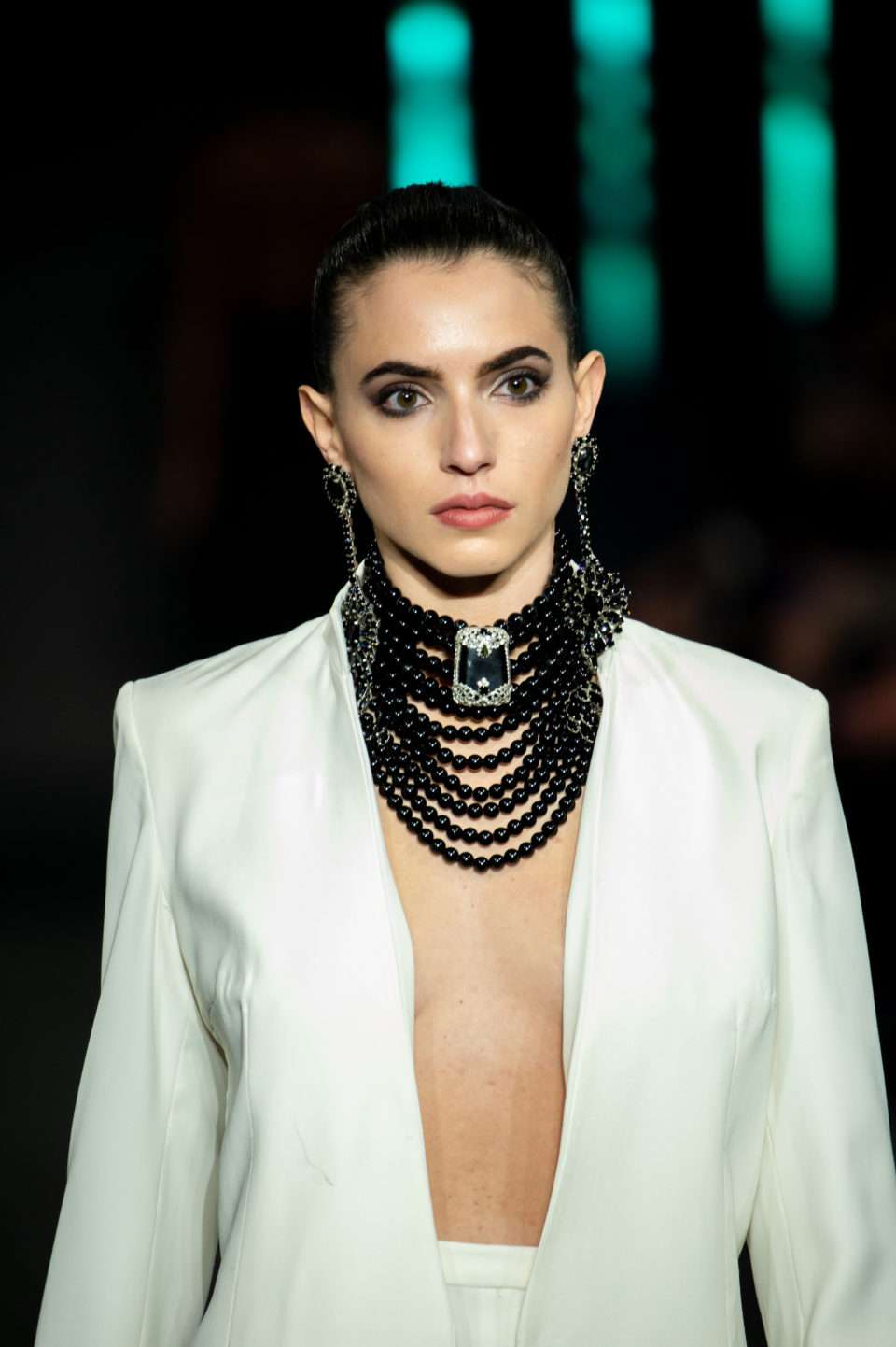 Female model wearing black statement earrings and a luxurious Baroqco necklace, styled with a white suit from Blue Valentine for Alta Roma 2024. The jewelry features Swarovski crystals, genuine CRYSTAL®, semi-precious stones, and freshwater pearls, set in recycled silver or gold plating—nickel-free. A true embodiment of luxury, haute couture, and regal elegance.